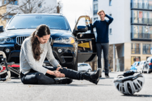 A woman sits on the ground beside a car, with her bicycle resting on the ground next to her.